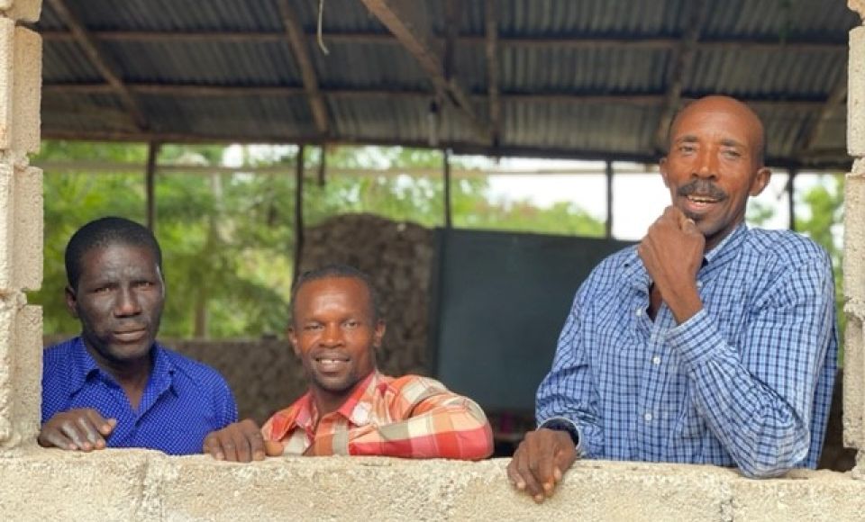 three men looking over a brick wall into the camera, smiling.
