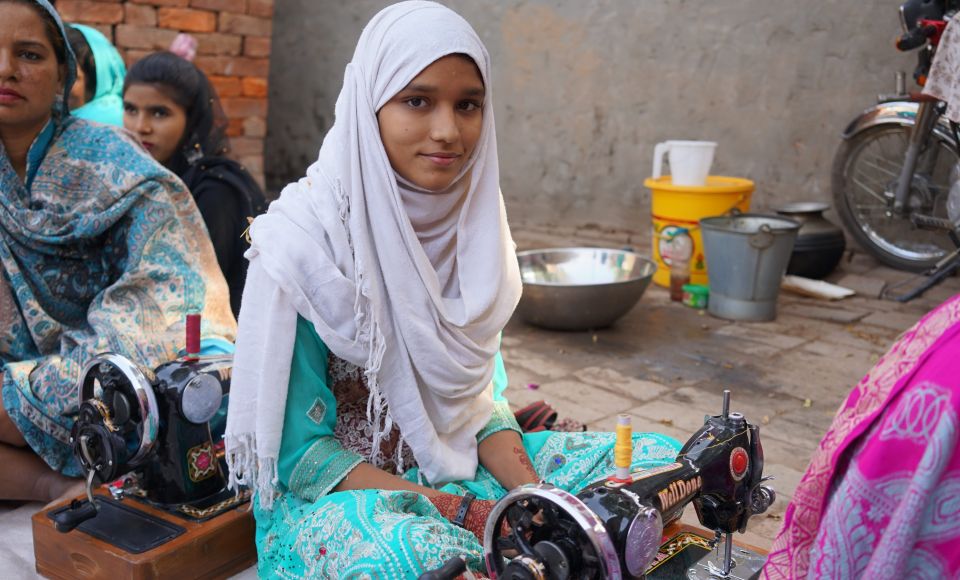 A young girl in a white headscarf and turquoise outfit sits at a sewing machine, with other women around her.