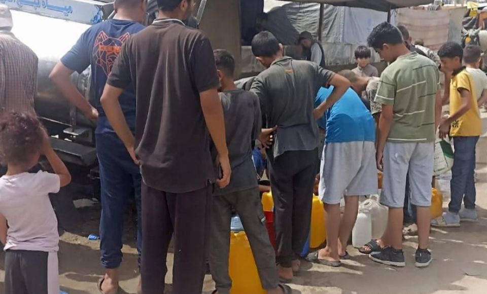  A group of people, including children and adults, are gathered around a water tanker, filling containers with water.