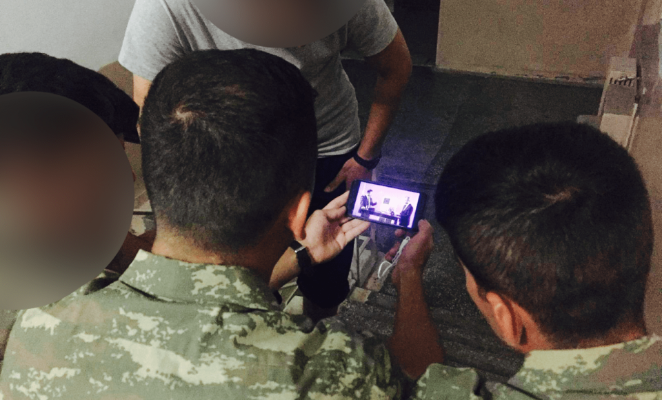 A group of army individuals huddled around a phone watching an interview.